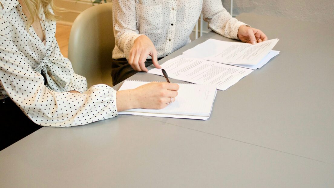 Duas mulheres analisando papéis e fazendo anotações em um caderno. Os rostos delas não aparecem. Elas podem estar procurando portabilidade de crédito